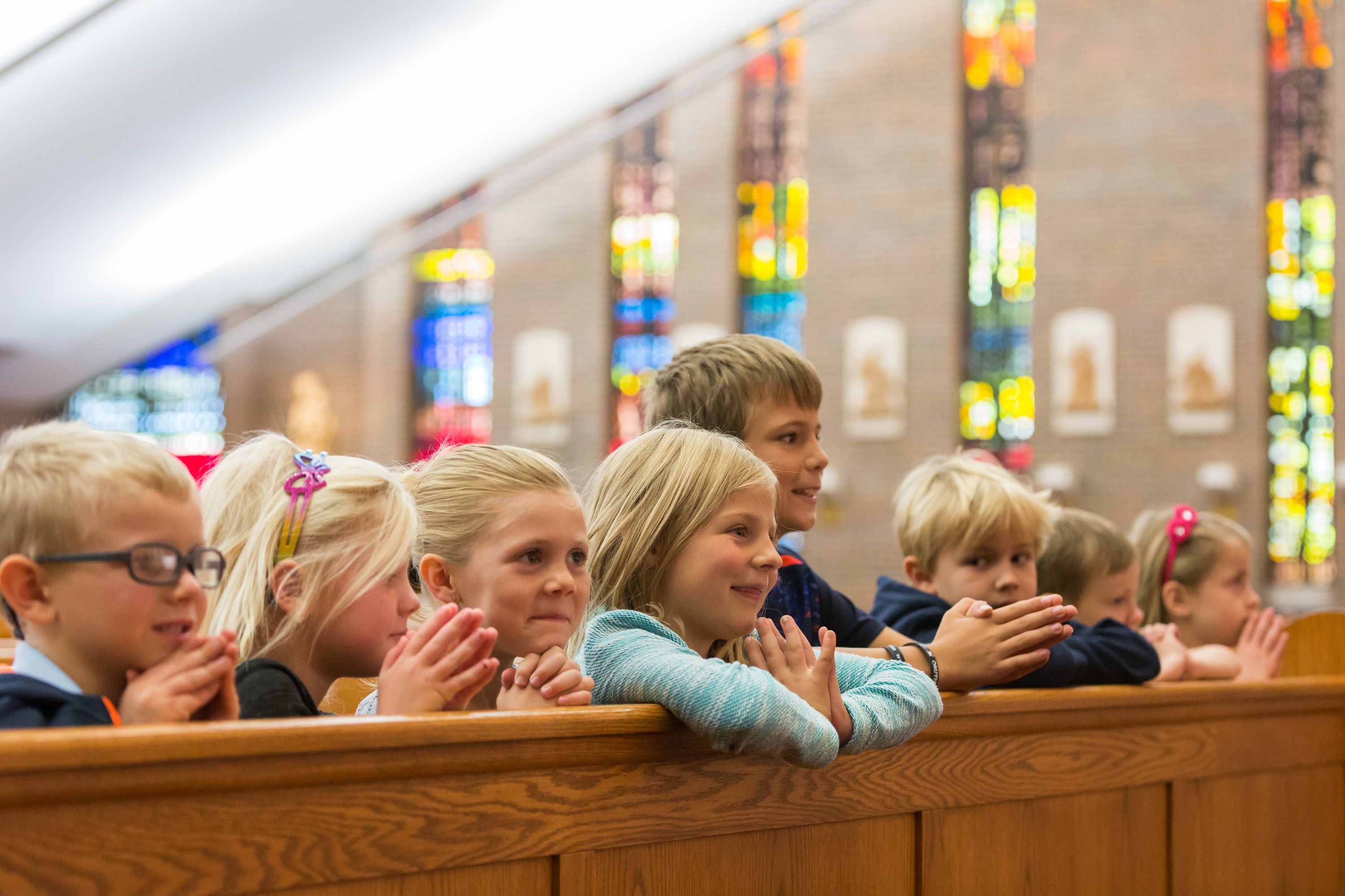 Young students kneel with folded hands in a church pew beneath stained-glass windows.