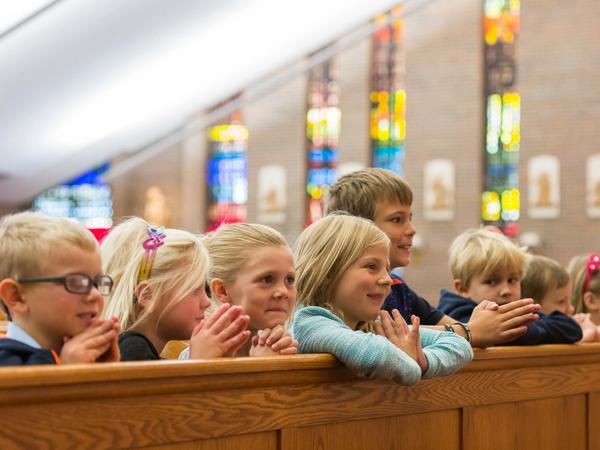 Young students kneel with folded hands in a church pew beneath stained-glass windows.