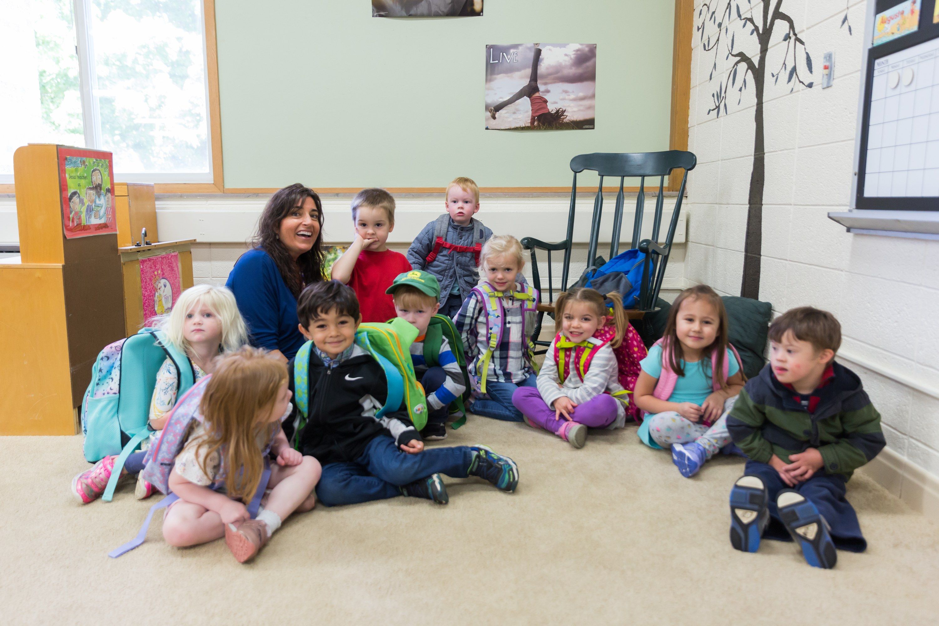 Preschoolers with their backpacks on sit together on the classroom rug with their smiling teacher.