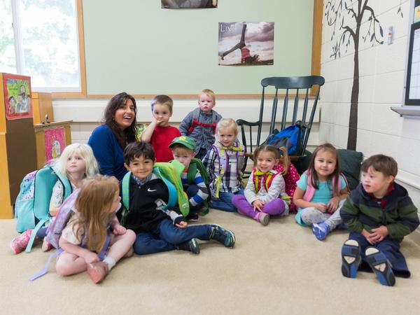 Preschoolers with their backpacks on sit together on the classroom rug with their smiling teacher.