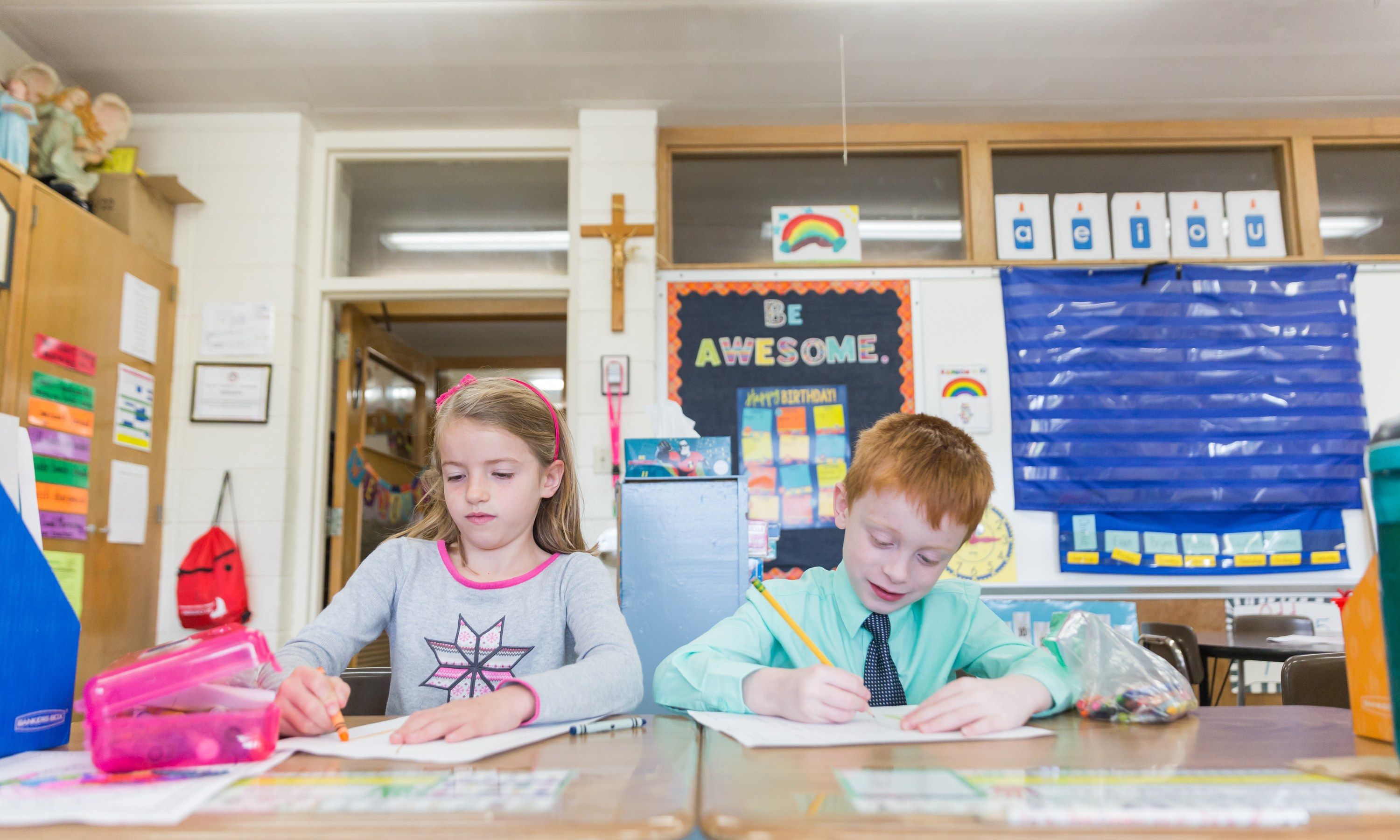 Two first graders write at their desks under a crucifix and a Be Awesome poster.