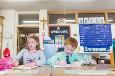 Two first graders write at their desks under a crucifix and a Be Awesome poster.