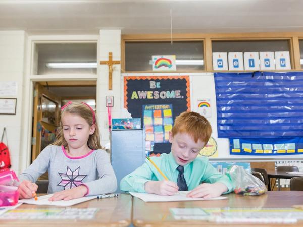 Two first graders write at their desks under a crucifix and a Be Awesome poster.