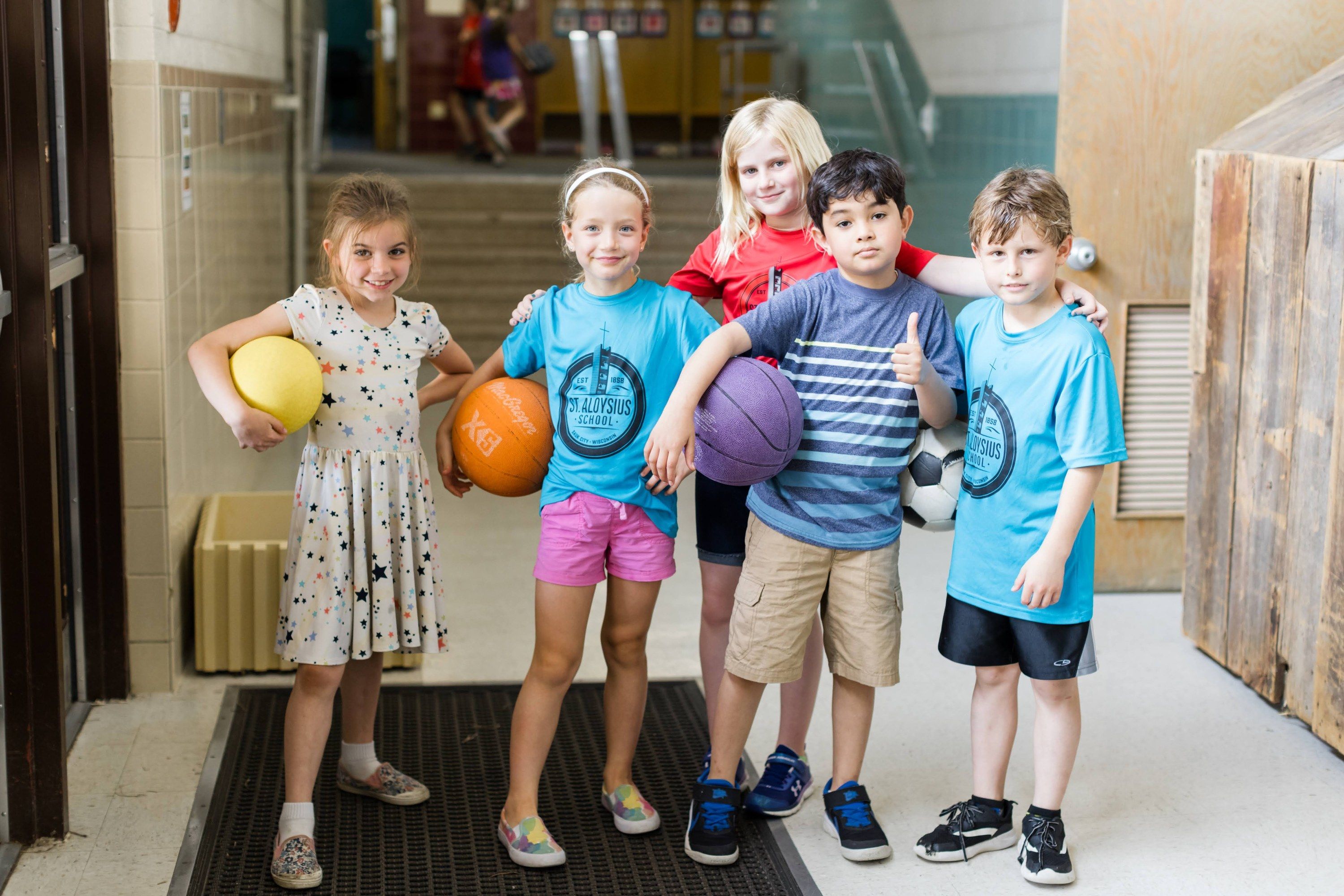 Five smiling students in St. Aloysius shirts hold playground balls in a school hallway.