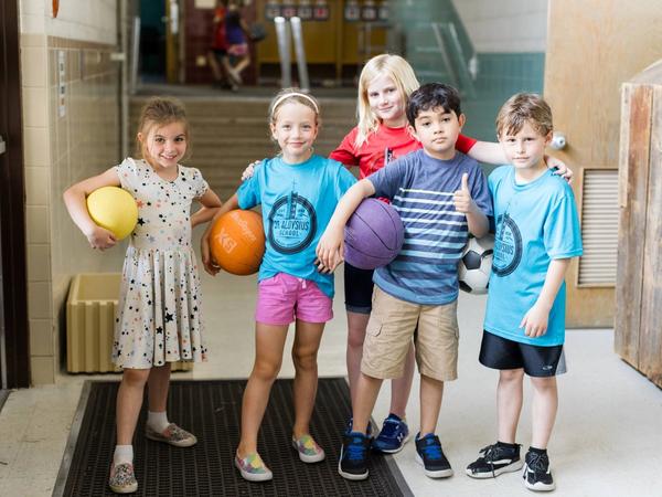 Five smiling students in St. Aloysius shirts hold playground balls in a school hallway.