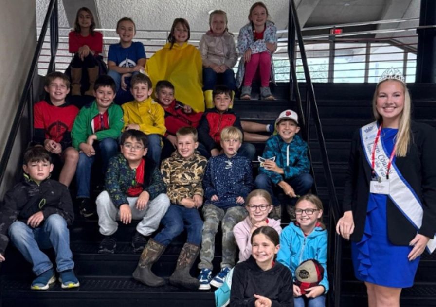 Fourth and fifth graders posing on a staircase with a young woman wearing a sash and tiara.