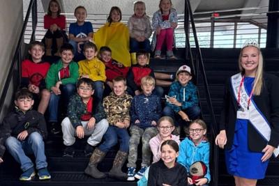 Fourth and fifth graders posing on a staircase with a young woman wearing a sash and tiara.