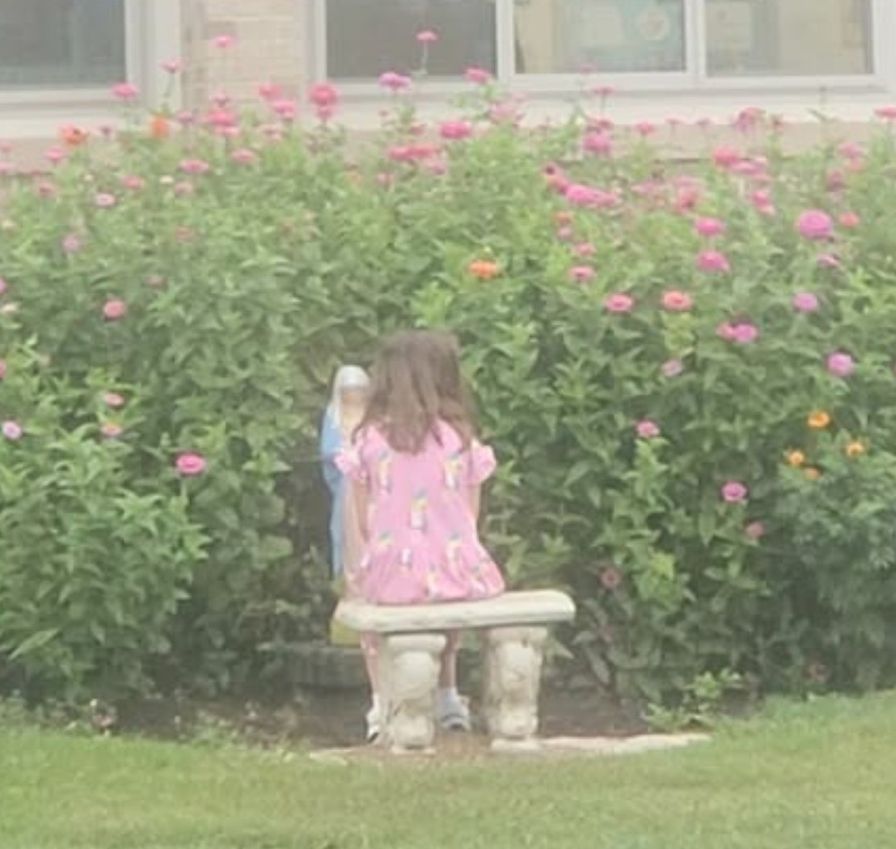 A student sitting on a bench in the flowering Mary Garden at recess.