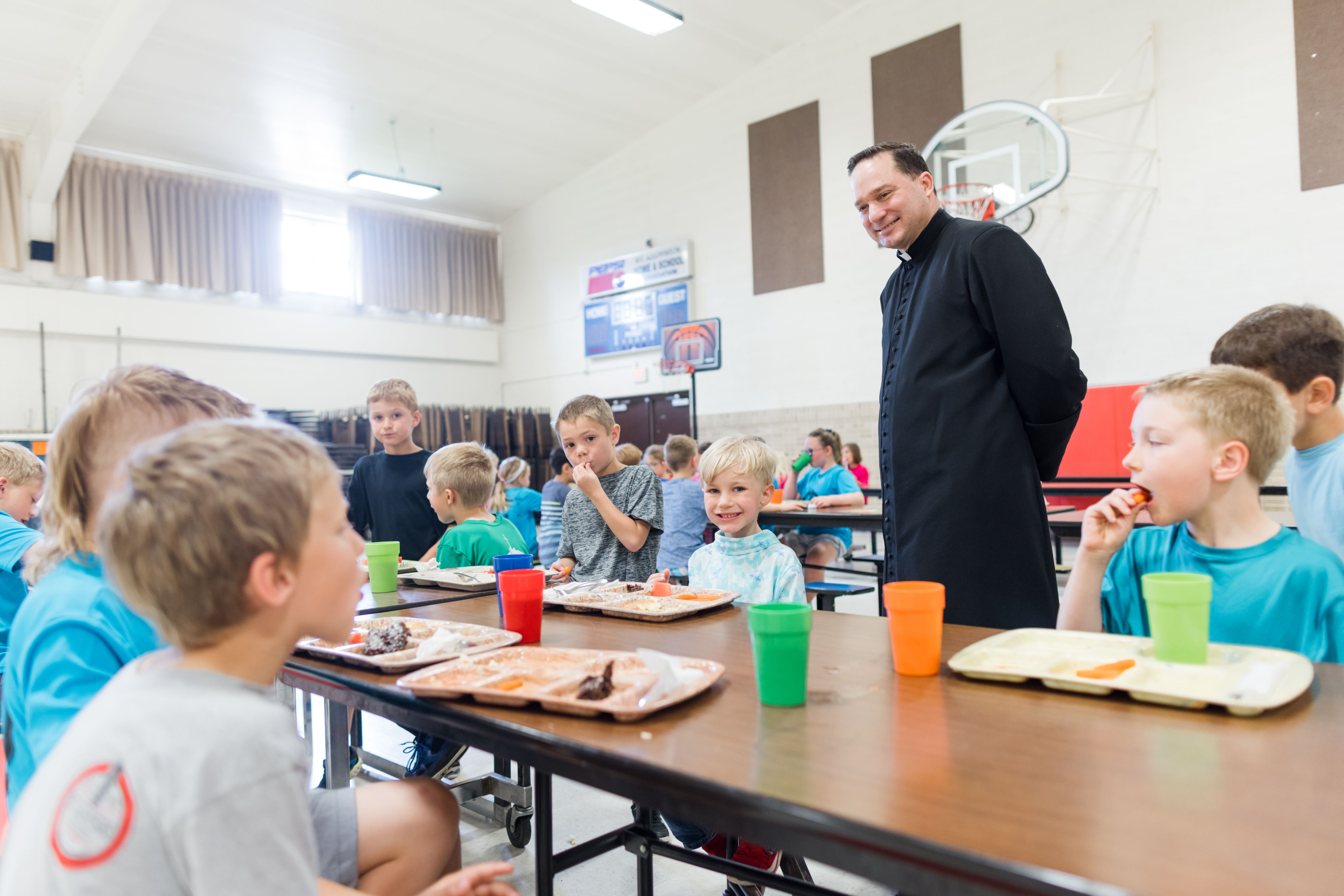 Students eat lunch together in the school gym while a priest visits their table.