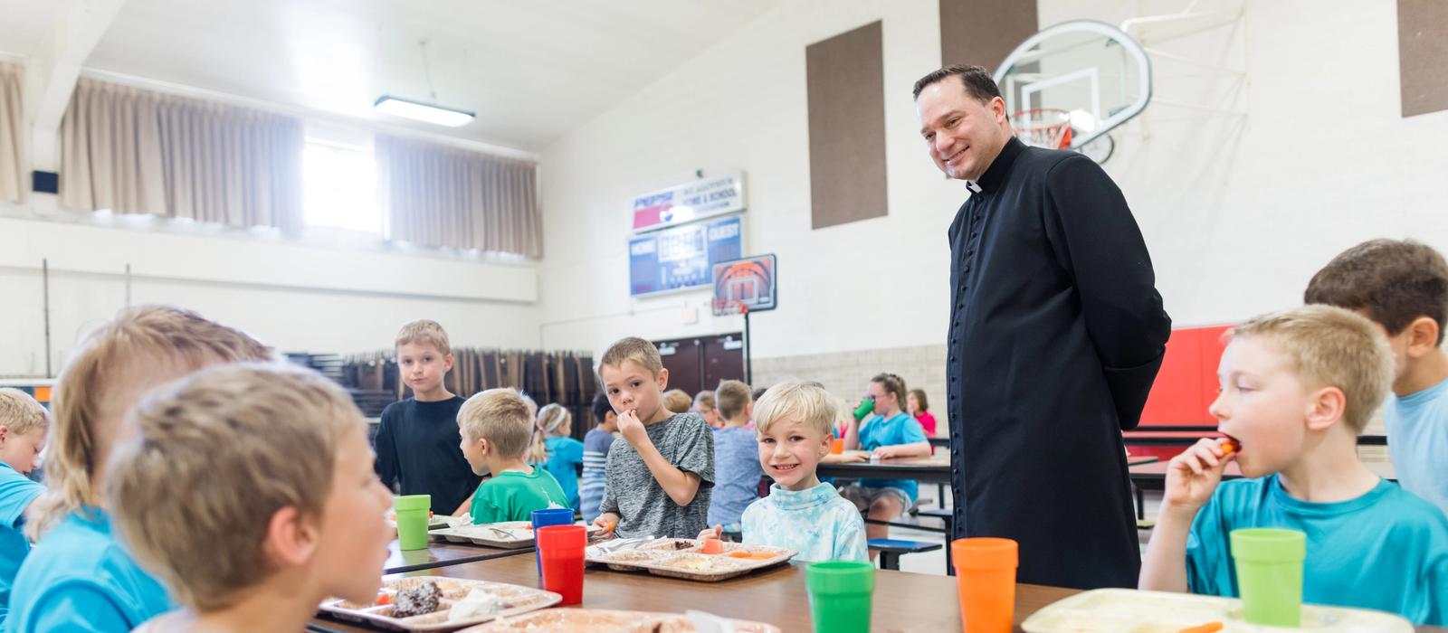 Students eat lunch together in the school gym while a priest visits their table.