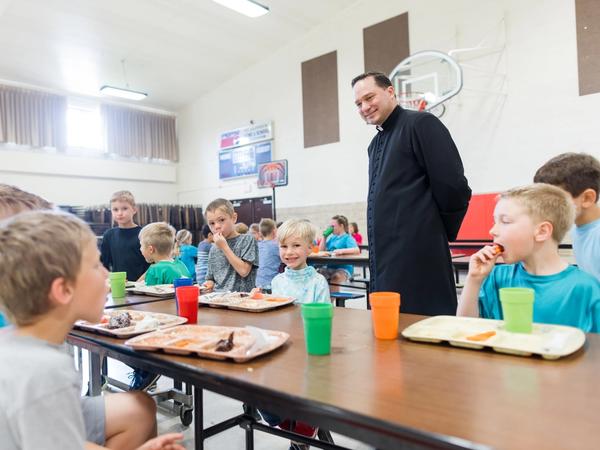 Students eat lunch together in the school gym while a priest visits their table.