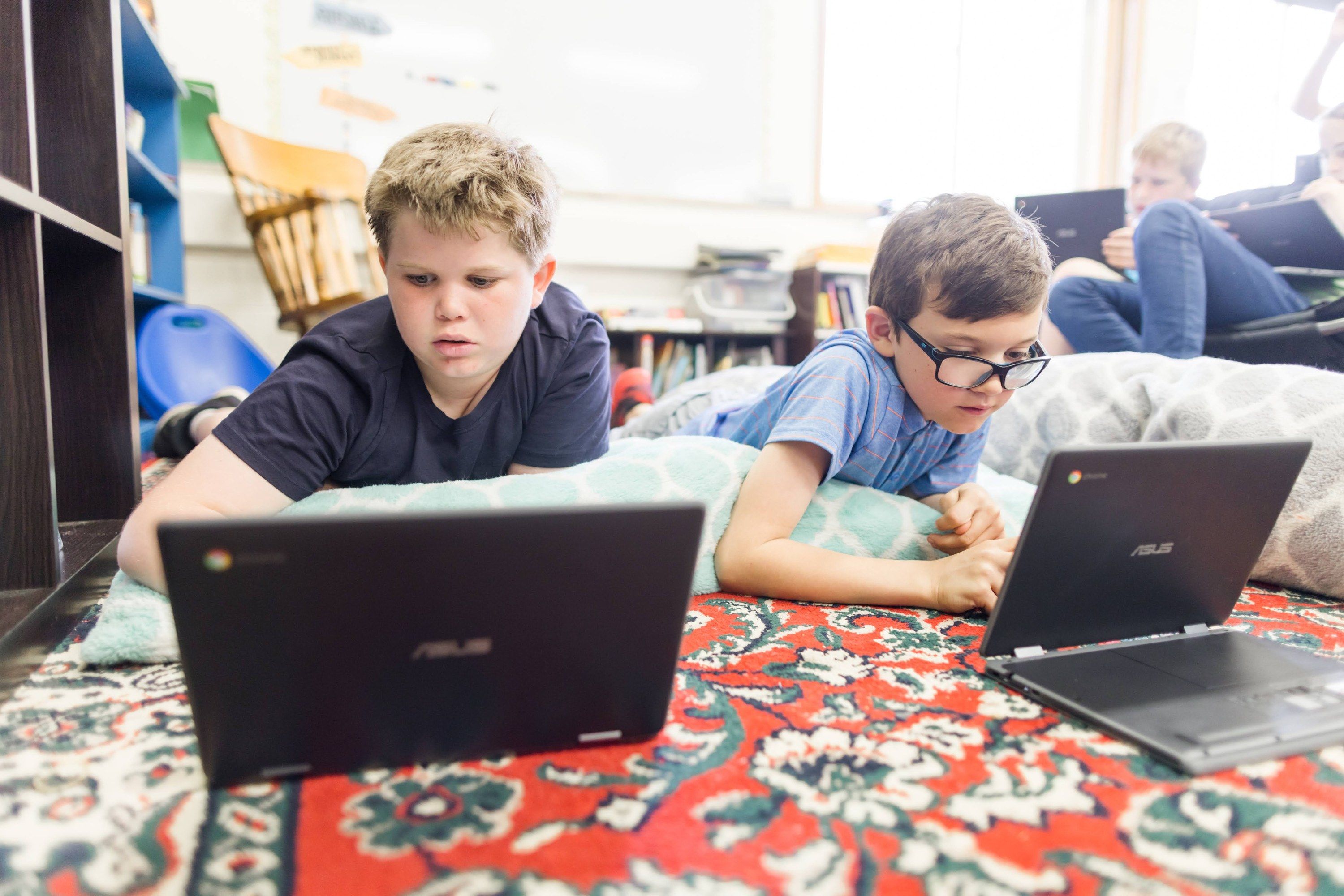 Two students lie on a classroom rug working on Chromebooks.