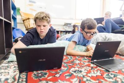 Two students lie on a classroom rug working on Chromebooks.