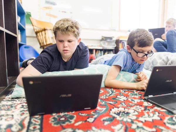 Two students lie on a classroom rug working on Chromebooks.
