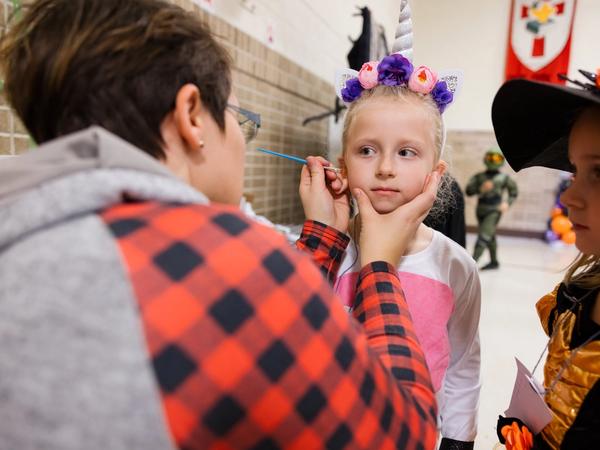 A volunteer paints a design on a young student's cheek at the all-school Halloween party.