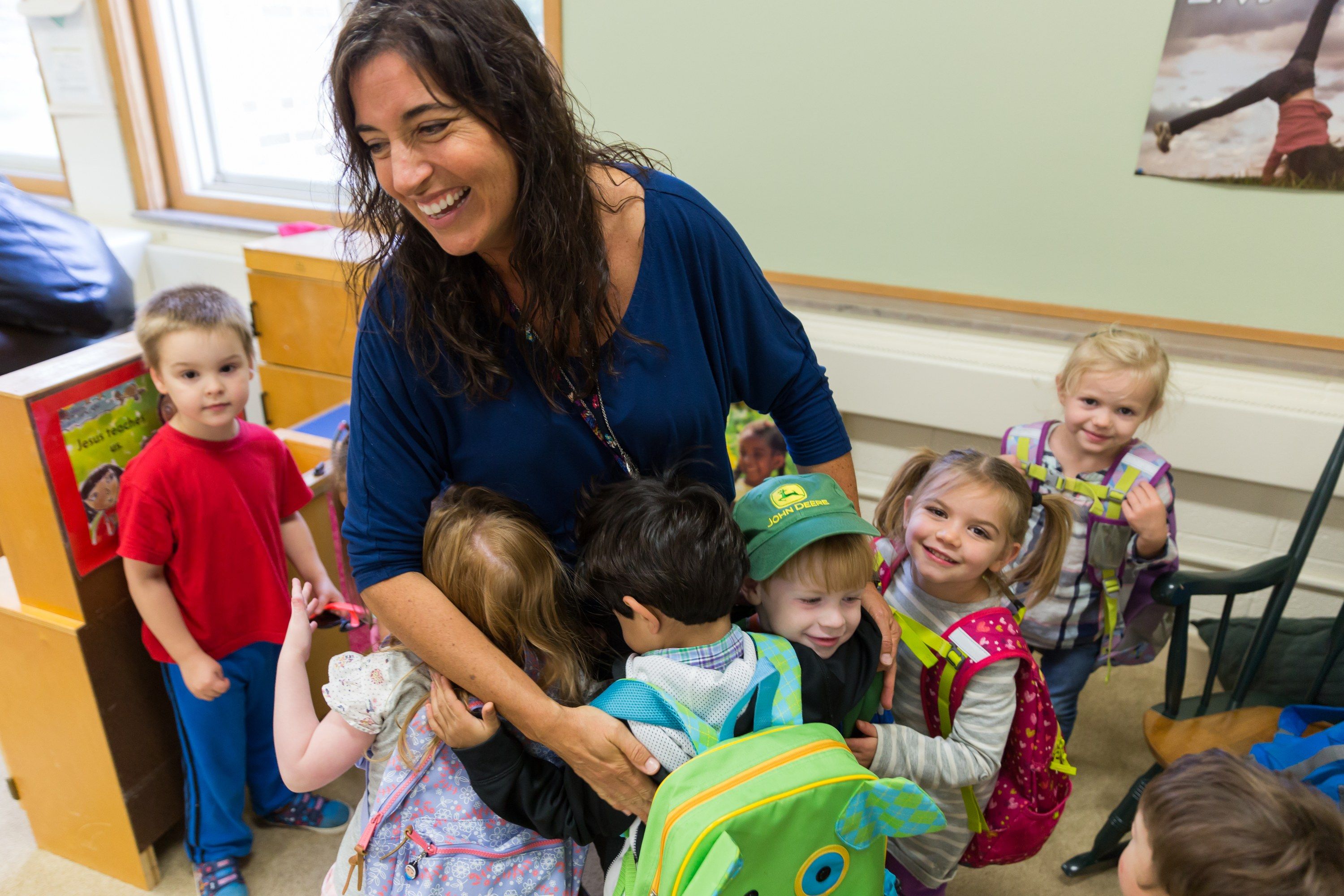 A smiling teacher wraps her preschool students, backpacks still on, in a group hug.