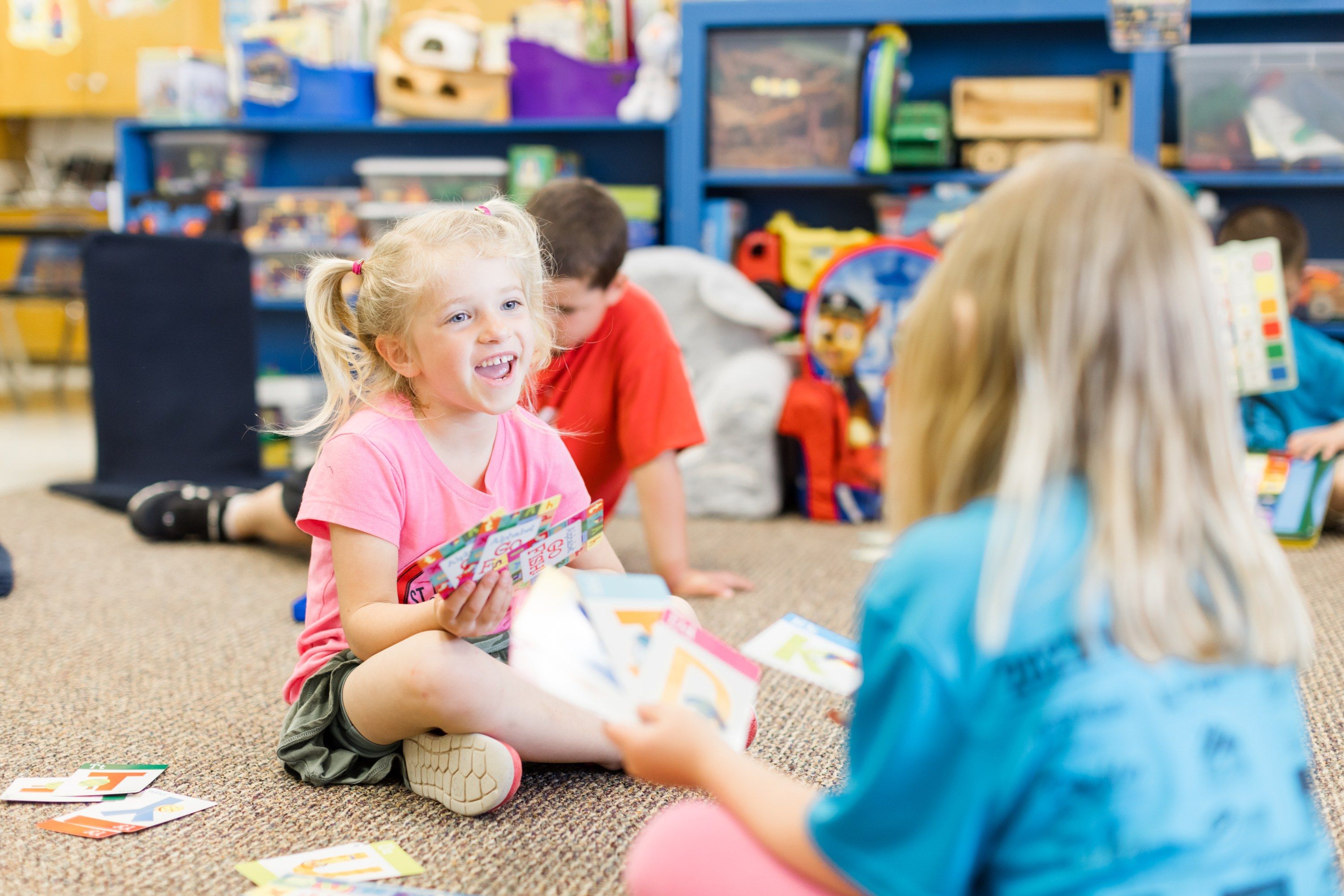 A kindergartner laughs while playing a card game with classmates on the classroom rug.
