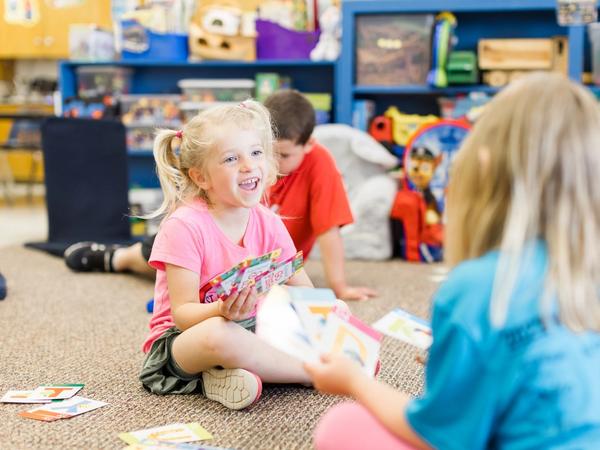 A kindergartner laughs while playing a card game with classmates on the classroom rug.