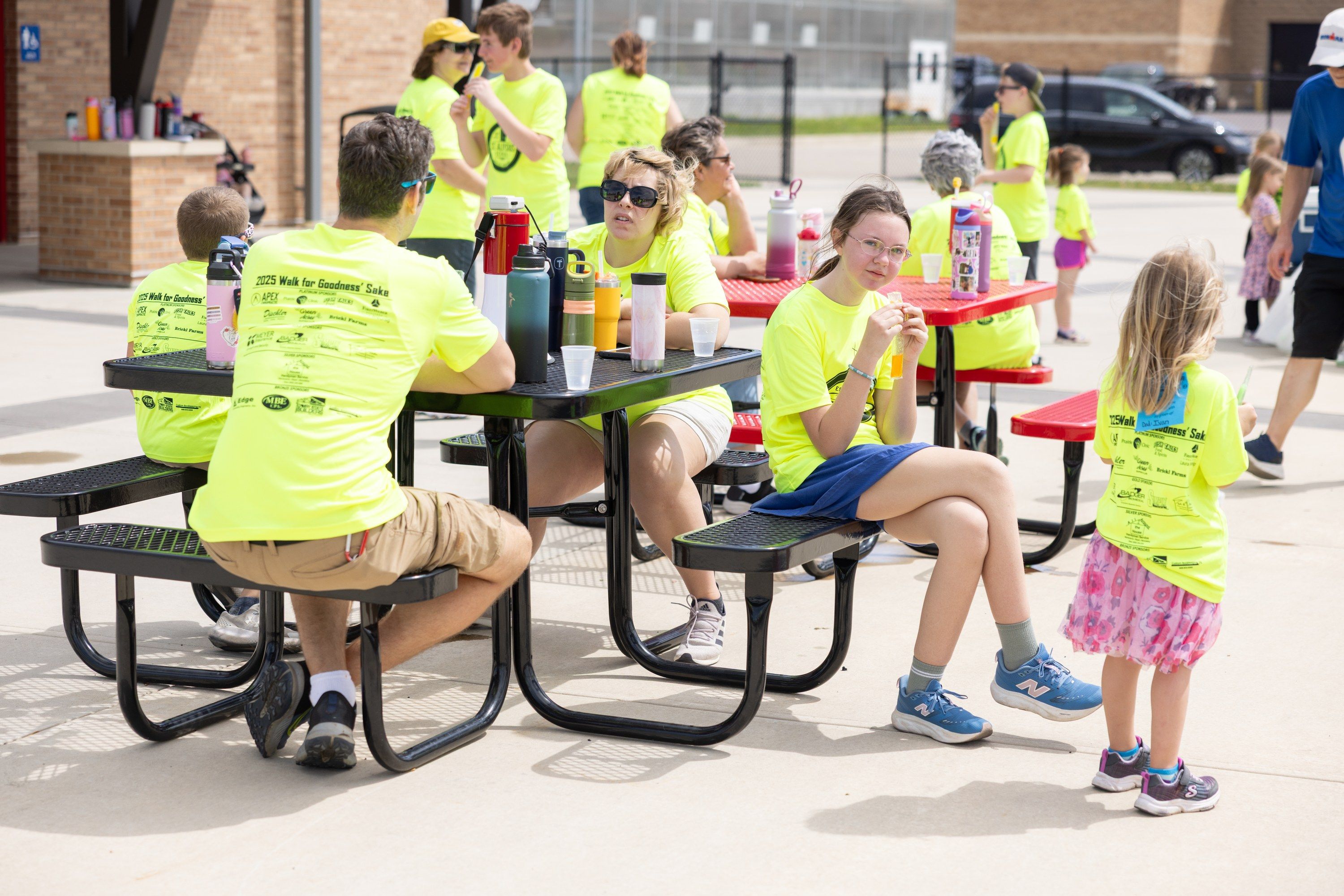 Students and adults in Walk for Goodness' Sake shirts take a break together at picnic tables.