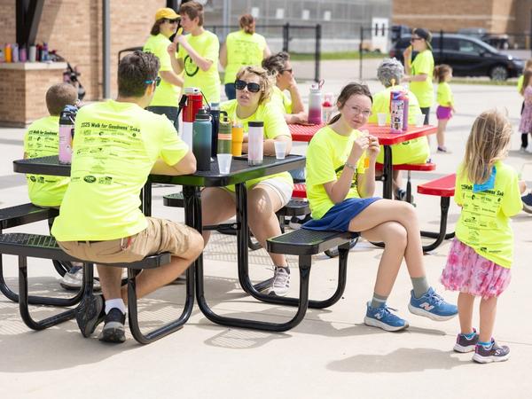 Students and adults in Walk for Goodness' Sake shirts take a break together at picnic tables.