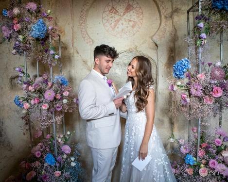 Braut und Bräutigam stehen in einem mit Blumen geschmückten sandsteinfarbenen Raum und lesen einander das Ehegelübde vor.