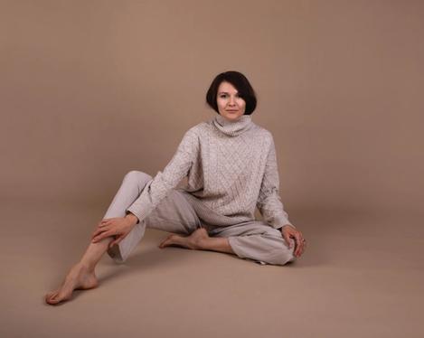 Dark-haired woman sits on the floor in beige trousers and turtleneck jumper in studio set of same colour. One leg is bent, the other stretched out. Her gaze is directed towards the camera.
