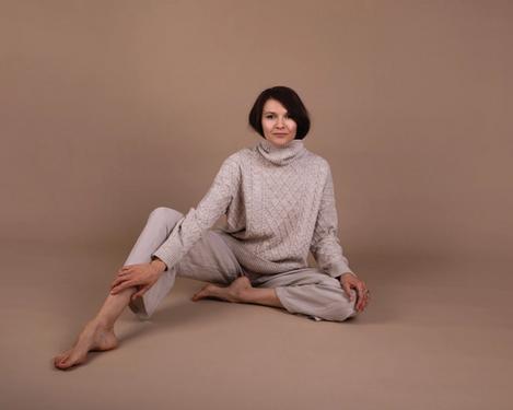 Dark-haired woman sits on the floor in beige trousers and turtleneck jumper in studio set of same colour. One leg is bent, the other stretched out. Her gaze is directed towards the camera.