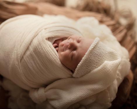 Baby smiling in sleep, wrapped in light cloths with light cap, lying in dark wooden bowl