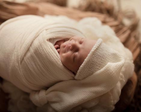 Im Schlaf lächelndes Baby, eingewickelt in helle Tücher mit heller Mütze, in dunkler Holzschale liegend
