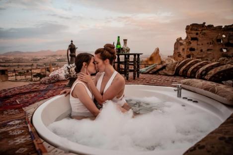 A same-sex female couple kiss each other in an outdoor bathtub. Stone ruins can be seen in the background.