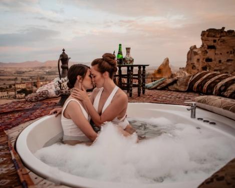 A same-sex female couple kiss each other in an outdoor bathtub. Stone ruins can be seen in the background.