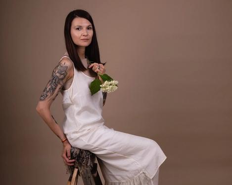 Dunkelhaarige Frau sitzt in hellem Kleid und in beigefarbenen Studioset auf einer Leiter. Sie hat eine Blume in der Hand und blickt in die Kamera.  