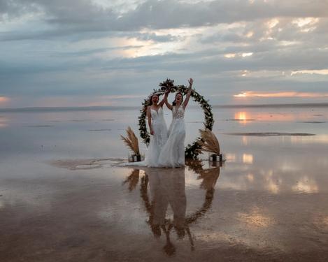 Same-sex female bride and groom standing side by side in front of an arch of flowers in a reflecting lake at sunset. They are happily holding their bridal bouquet aloft.