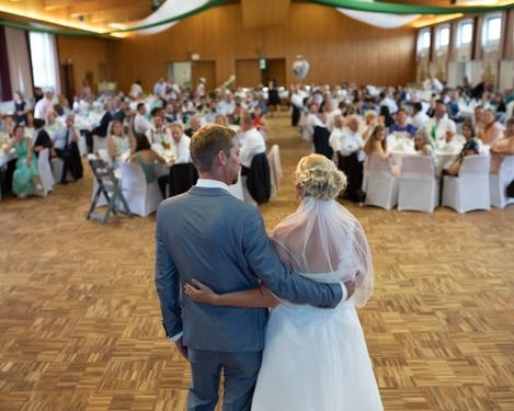 Braut und Bräutigam stehen mit dem Rücken zur Kamera und sprechen zu den Hochzeitsgästen im Saal