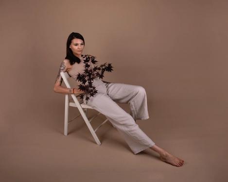 Dark-haired woman sits on an armchair in beige trousers and tight top in studio set of same colour. One leg is bent, the other stretched out. Her gaze wanders thoughtfully into the distance.
