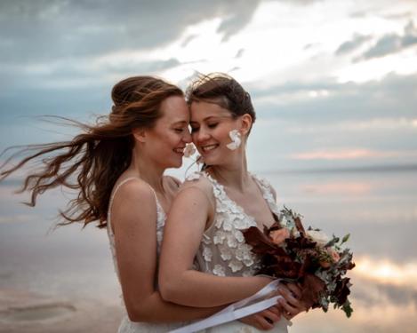 Gleichgeschlechtliches weibliches Brautpaar stehen hintereinander und seien einander lächelnd an. Die Haare wehen im Wind bei Sonnenuntergangsstimmung.