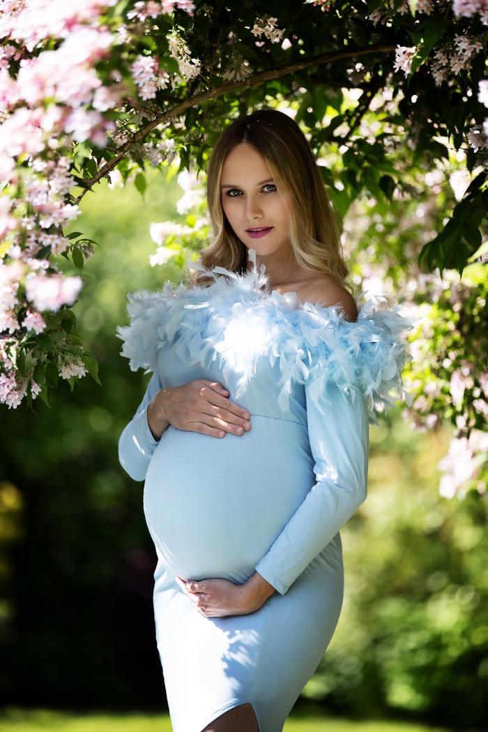 Schwangere Frau in hellblauem Kleid mit Federbesatz vor blühender Hecke