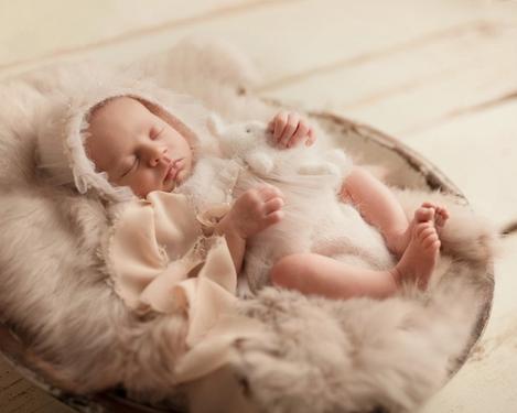 Baby in lace dress and bonnet in white wooden bowl padded with fur on light wooden floor