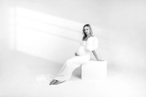 Pregnant woman sitting on white stool in white fitted dress and gathered sleeves on white background