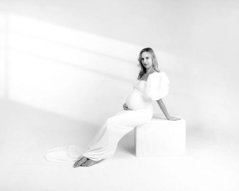 Schwangere Frau sitzt auf weißem Hocker in weiß anliegendem Kleid und gerafften Ärmeln vor weißem Hintergrund