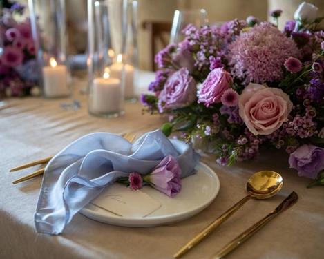 Ausschnitt der Hochzeitstafel mit rosa/lila Blumen, goldenem Besteck und hellblauer Serviette.