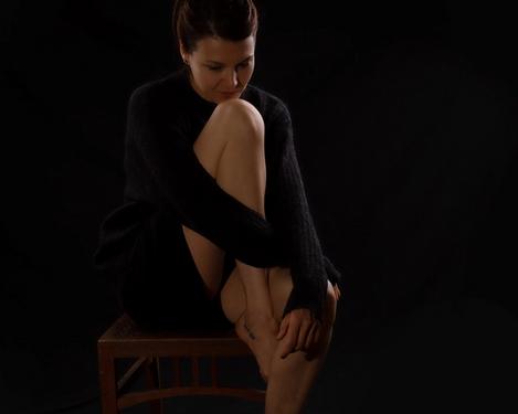 Schwarz gekleidete Frau sitzt auf einem Sessel, Kinn am Knie gestützt mit Blick nach unten