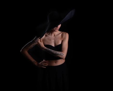 Portrait of a woman with a black hat and tattoos on her arm looking to the side. The face is half covered by the brim of the hat, black background.