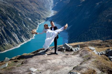 Groom holds bride in his hands, behind them a turquoise lake framed by mountains