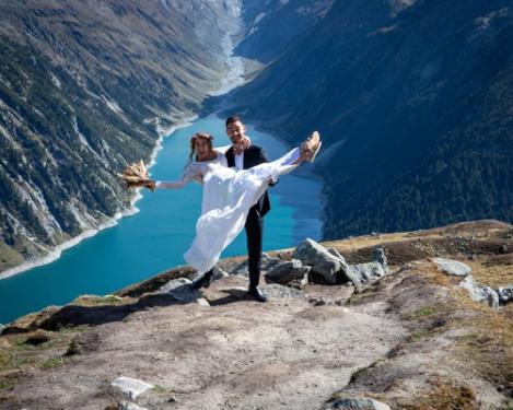 Groom holds bride in his hands, behind them a turquoise lake framed by mountains