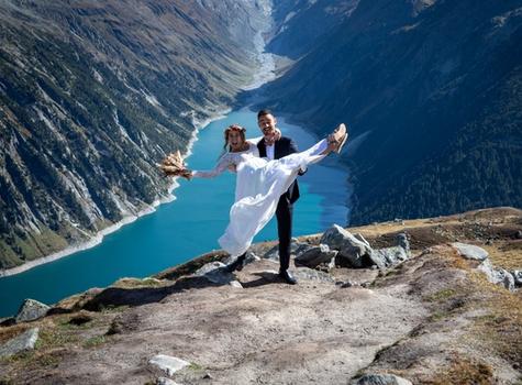 Hochzeitspaar in klassischer Kleidung in den Bergen. Im Hintergrund sind Berge und ein türkisblauer Stausee zu sehen. 