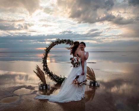 Same-sex female bride and groom standing in front of an arch of flowers in a reflecting lake at sunset.