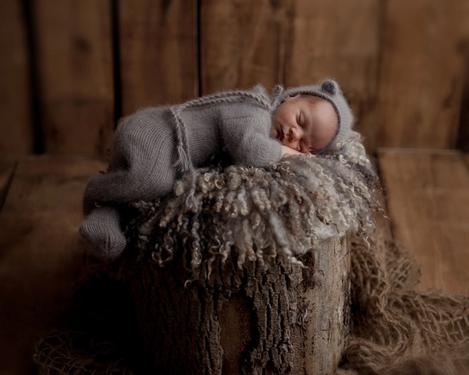 Newborn in brown jumpsuit sleeps on a tree trunk padded with fur.
