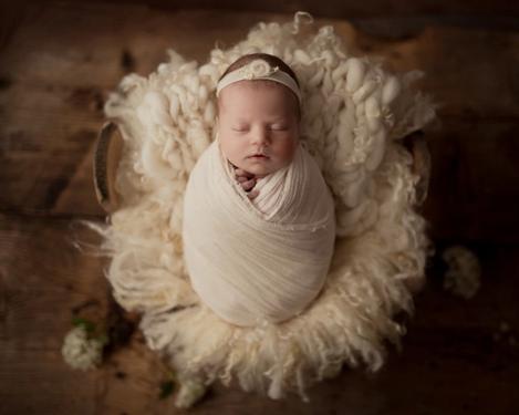 Baby mit Stirnband eingewickelt in ein creme farbiges Tuch auf braunem Holzboden