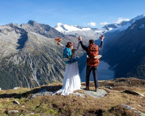 Rückenansicht eines Brautpaares mit Rucksack in den Bergen mit Sicht auf einen Stausee